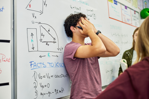 Teenager stressed out from school and relationships - image of teenage boy with marker in his hand in front of complicated math equations, he has his hands covering his face, while 2 teenage girls stand nearby.