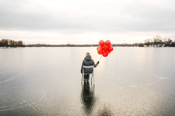 Imagery of emotional numbness - woman alone on a large frozen lake, sitting in a chair with a red balloon bouquet, staring off into the distance.