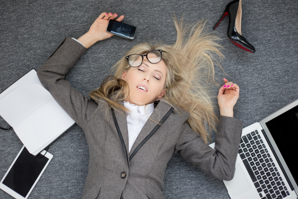 Professional woman exhuasted from burnout. Images shows woman laying on floor with hair askew, eyes closed, and surrounded by phone, laptop, ipad, notebook, and stiletto show
