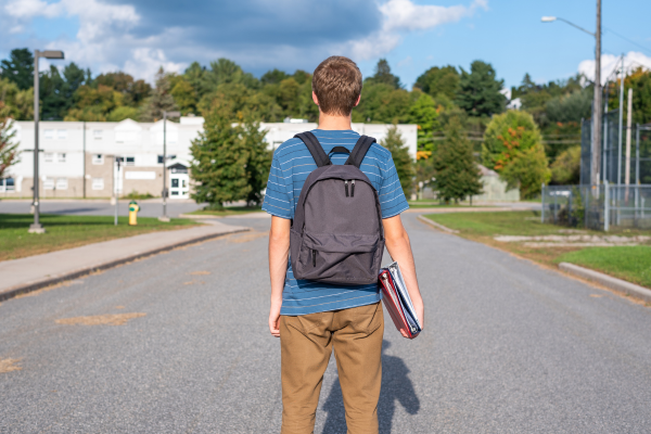 Teen wearing backpack and holding books while standing in the street looking at his school.